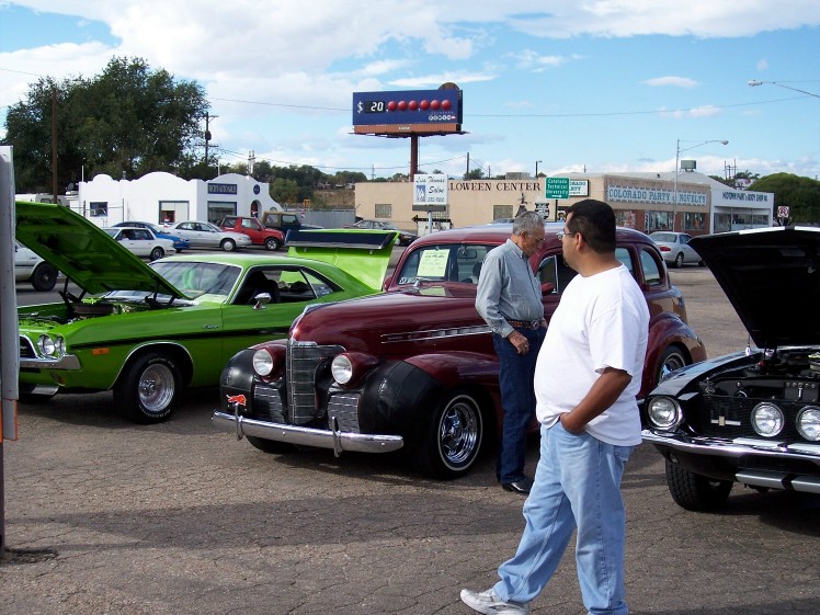 Several classic cars are parked outside a building, with people looking at them. A green muscle car has its hood up