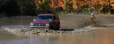 1979 Ford Bronco - A red SUV navigates a muddy obstacle course with a person on an ATV in the background.
