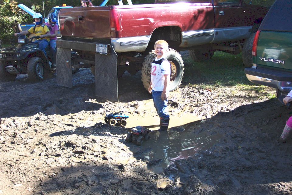 Kid in mud puddle with toy trucks, big red truck, green Yukon, and ATV in background