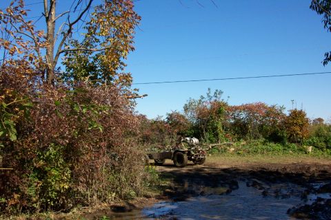 1979 Ford Bronco - An all-terrain vehicle pulls a trailer along a muddy track surrounded by autumn foliage under a clear blue sky.