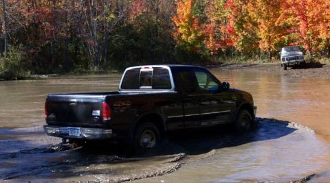 1979 Ford Bronco - A black pickup truck navigates through shallow, muddy water with autumn foliage in the background.