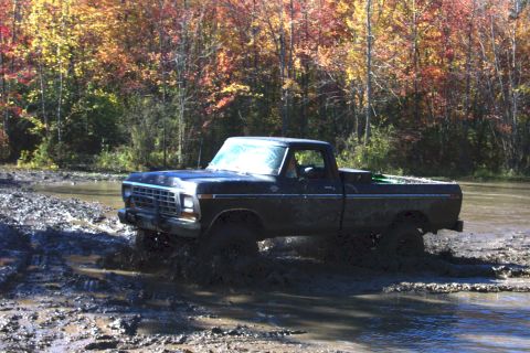 1979 Ford Bronco - A dark blue pickup truck, likely from the late 1970s, navigates through a muddy terrain with colorful autumn trees in the background.