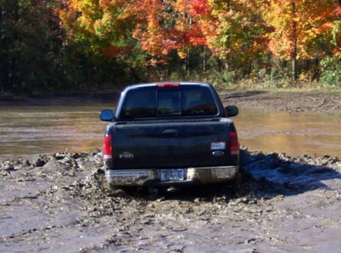 1979 Ford Bronco - A Ford F-150 is stuck in the mud near a body of water, with autumn trees in the background.