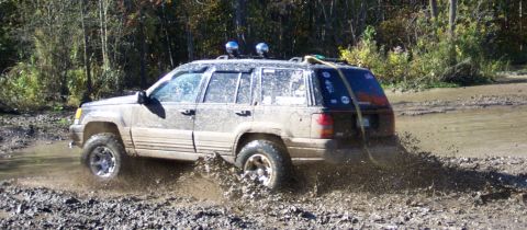 1979 Ford Bronco - A Jeep Grand Cherokee navigates a muddy off-road course, kicking up a large spray of water and dirt.