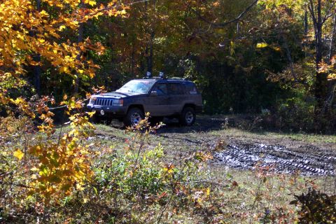 1979 Ford Bronco - Jeep Grand Cherokee navigating a muddy path during autumn, surrounded by trees with yellow leaves.