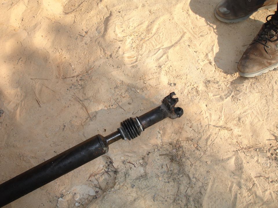 Driveshaft with a broken U-joint resting on sandy ground, with two pairs of boots visible in the upper right