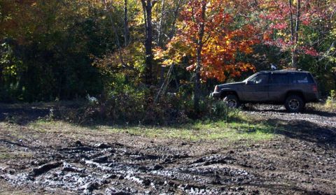 1979 Ford Bronco - A Jeep Grand Cherokee sits off-road on a muddy path surrounded by trees in autumn colors.