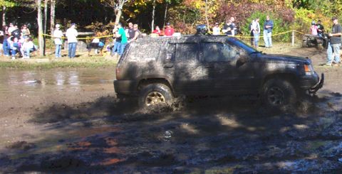 1979 Ford Bronco - A Jeep Grand Cherokee navigates a challenging muddy course, kicking up spray as onlookers watch.