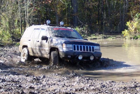 1979 Ford Bronco - This Jeep Grand Cherokee is navigating a muddy off-road course with trees and water visible.