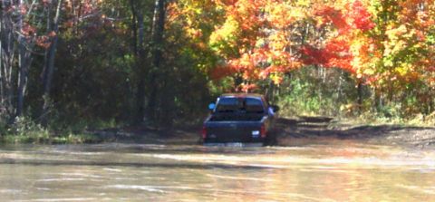 1979 Ford Bronco - Pickup truck navigates through a muddy water obstacle during autumn.