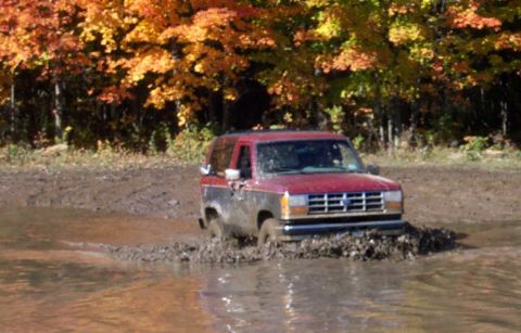 1979 Ford Bronco - A red and black SUV navigates a muddy obstacle course with fall foliage overhead.