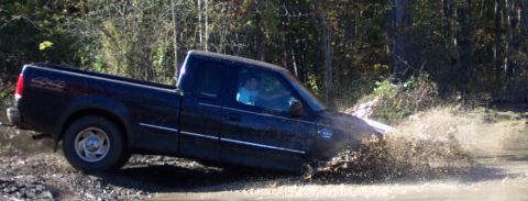 1979 Ford Bronco - Pickup truck driving through a muddy area with trees in the background.