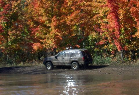 1979 Ford Bronco - A muddy SUV navigates through a shallow body of water, surrounded by vibrant autumn trees.
