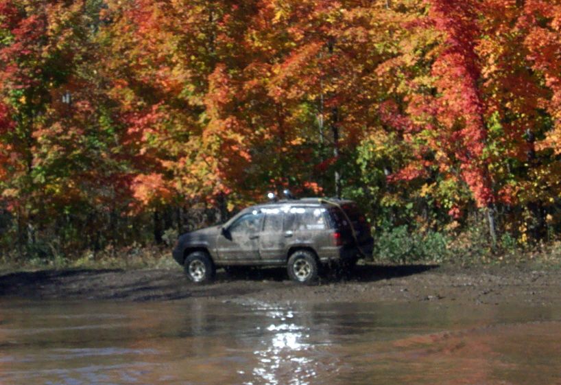 Muddy SUV driving through water with fall foliage in background