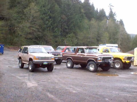 1976 Ford F-100 - Several Ford trucks and SUVs are parked in a gravel lot with trees in the background.