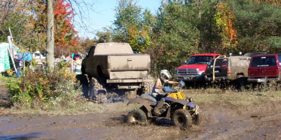 ATV splashing mud in front of a lifted pickup truck and other off-road vehicles in a muddy field