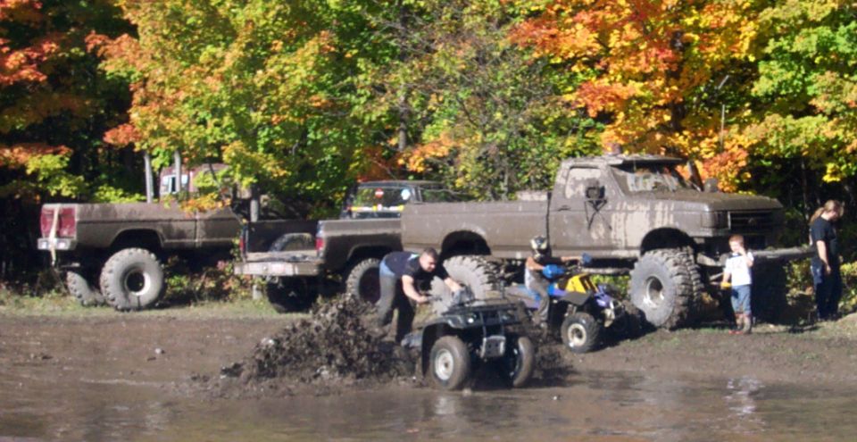 Several muddy pickup trucks and ATVs are in a wooded area with fall foliage. People are interacting with the vehicles in the mud