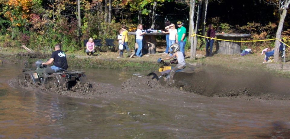 Two ATVs splashing through a muddy track with spectators watching from the bank