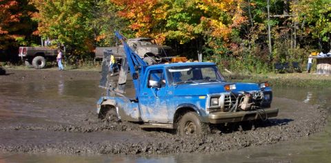 1979 Ford Bronco - A blue tow truck, appearing to be a 1979 Ford F-Series truck, navigates through a muddy off-road course with autumn foliage in the backgrou…