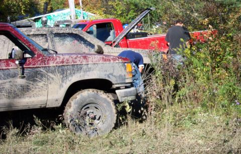 1979 Ford Bronco - Two off-road vehicles, a maroon SUV and a red pickup truck, are covered in mud in a grassy, wooded area.
