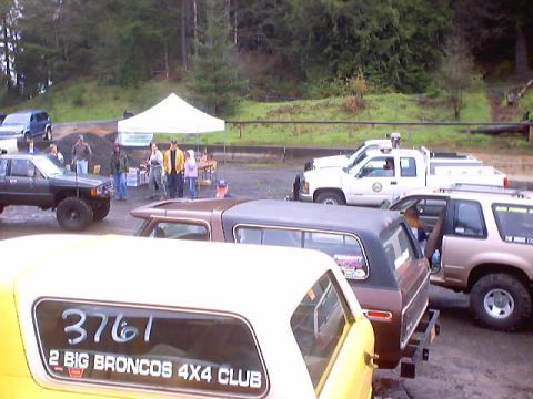 1976 Ford F-100 - A collection of trucks and SUVs are gathered in an outdoor lot, with one yellow vehicle prominently displaying club affiliation.