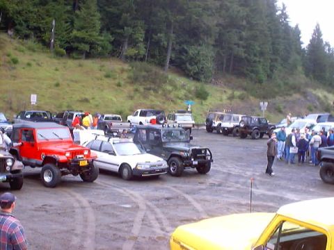 1976 Ford F-100 - Many off-road vehicles and people gathered in a parking area with a wooded hillside.