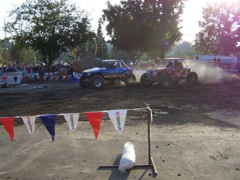 1995 Ford Bronco - Two custom off-road vehicles compete in a race, generating dust as they speed across the dirt track.