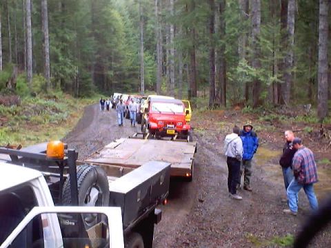 1976 Ford F-100 - A group of off-road vehicles are seen on a dirt road in a wooded area, with some people gathered near a trailer.