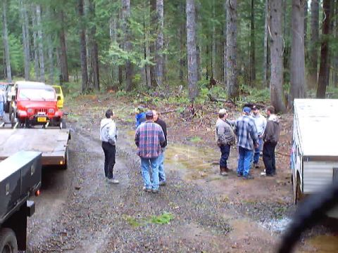 1976 Ford F-100 - A red Jeep Wrangler is loaded onto a trailer, with several people gathered nearby in a forest setting.