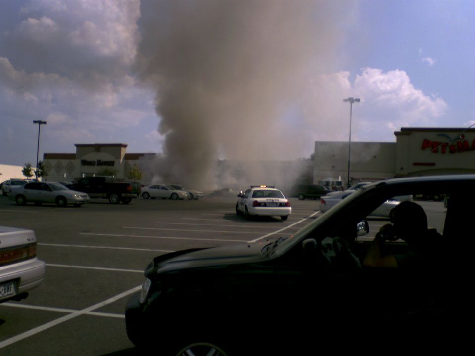 Parking lot with a large smoke plume rising from behind buildings, a police car is visible