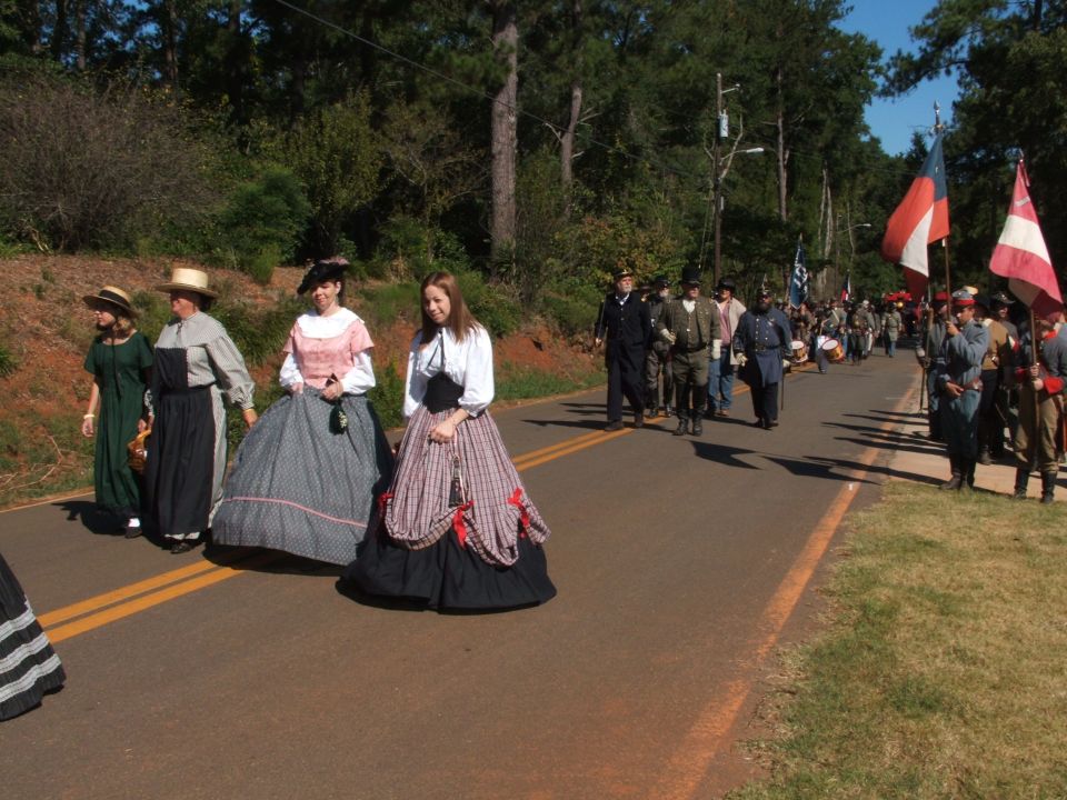 People in 19th-century clothing march on a paved road with trees and grass on the sides