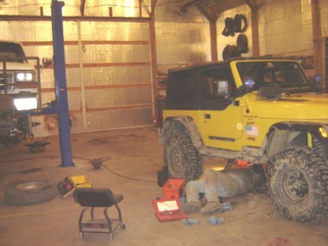 1992 Ford Bronco - A mechanic works under a yellow Jeep Wrangler in a garage, with another vehicle on a lift in the background.