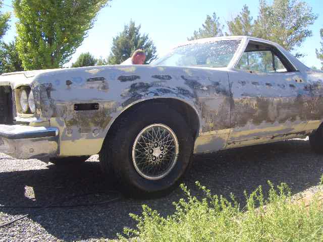 Ford Ranchero in gray primer with stripped paint patches, parked on gravel, front three-quarter view