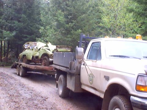 1976 Ford F-100 - A flatbed tow truck transports a heavily damaged vehicle on a trailer through a wooded area.