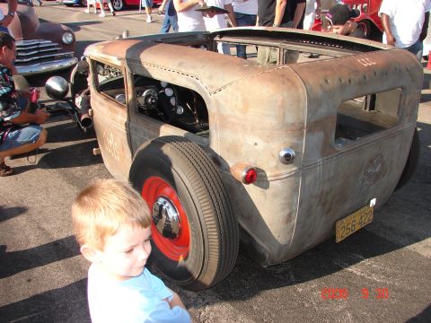 1987 Ford Bronco - A young boy stands next to a weathered rat rod at an outdoor car show.