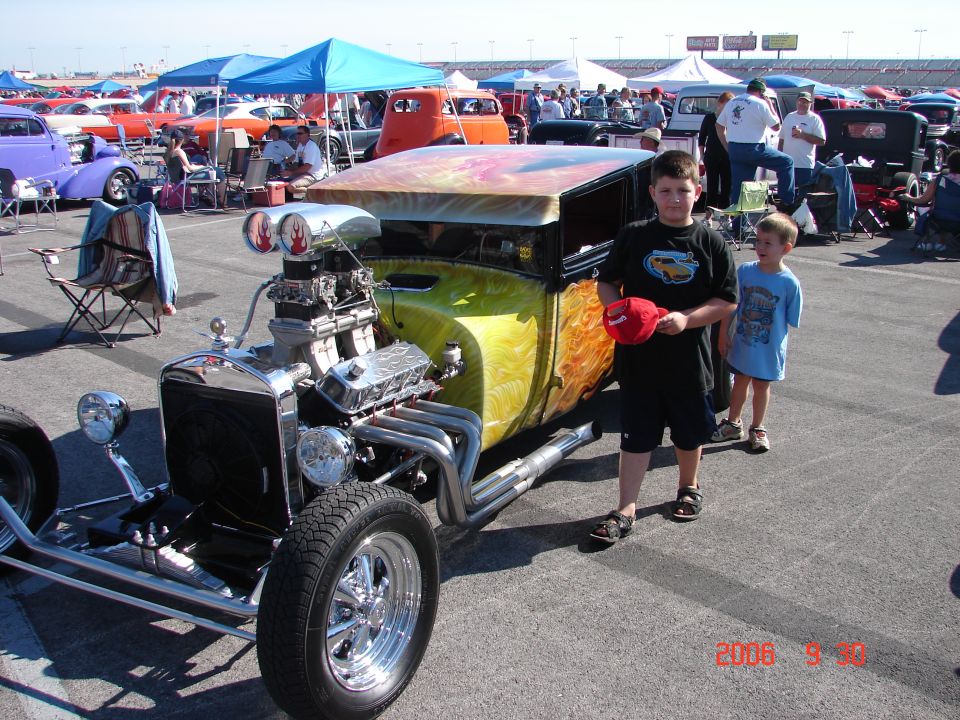 Custom hot rod with exposed engine and flame paint, two boys standing nearby. Other cars and tents in background