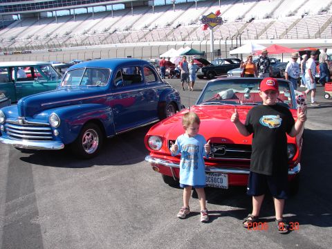 1987 Ford Bronco - Boys pose with classic cars at a race track event, with grandstands and sponsor signs visible.
