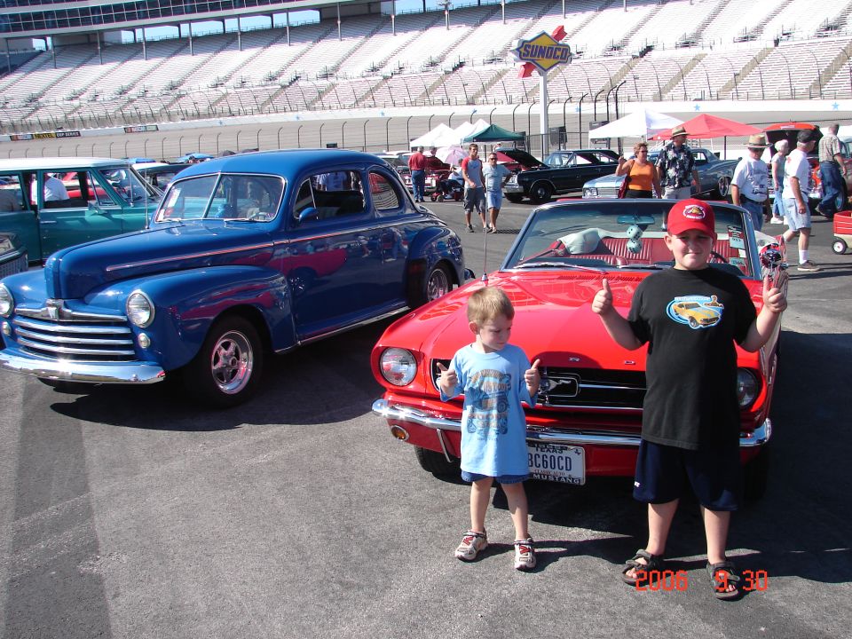 Two boys give thumbs up in front of a blue classic car and a red classic car at a race track with Sunoco signage