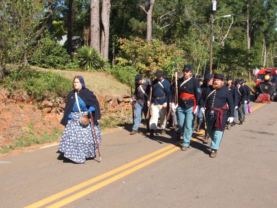 Civil War reenactors in uniform marching down a road, one woman in period dress walks ahead