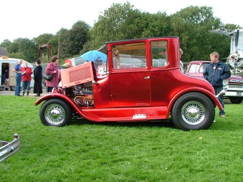 2001 Ford Fiesta - A classic red hot rod coupe is displayed on a grassy field with several people observing it.