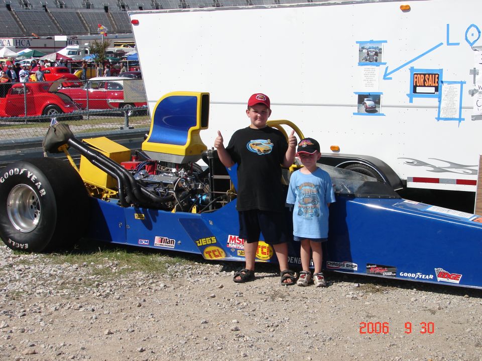 Two boys stand next to a blue and yellow dragster with a large rear tire. A white trailer is behind them