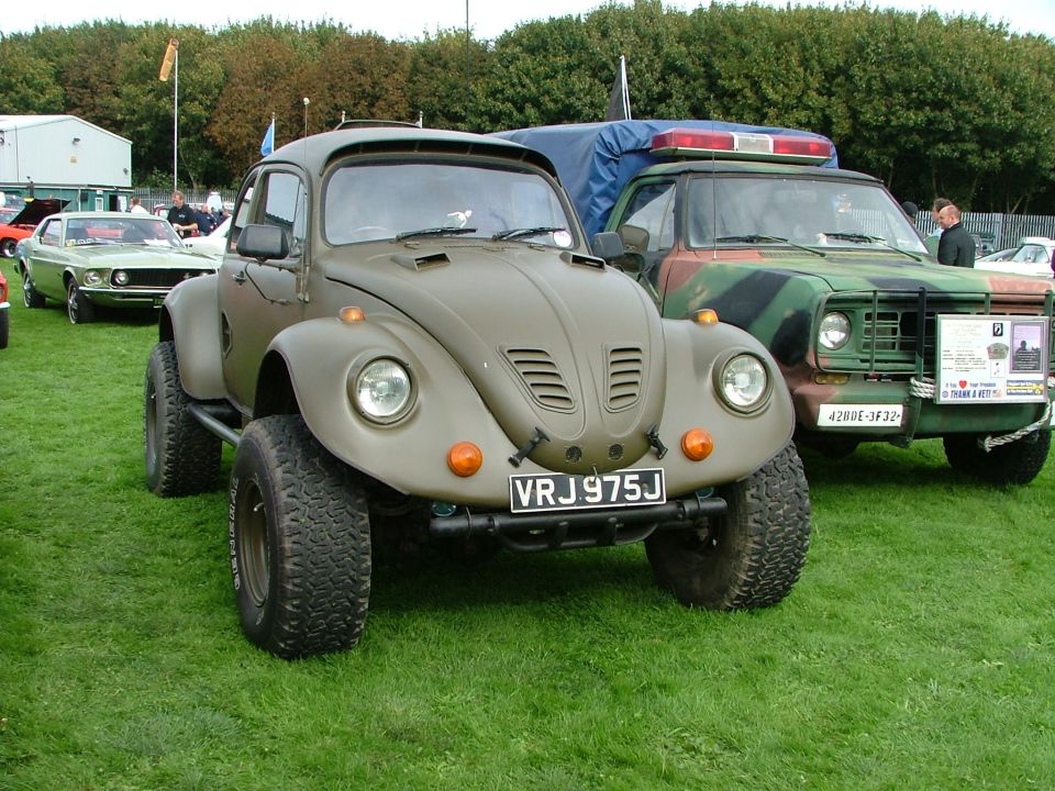 Olive green modified VW Beetle with large tires and a camo truck behind it