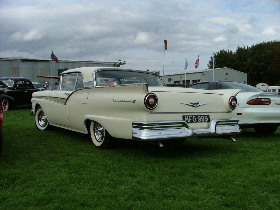 Rear view of a cream 1957 Ford Fairlane with a license plate MFO 999. A white Camaro is parked behind it