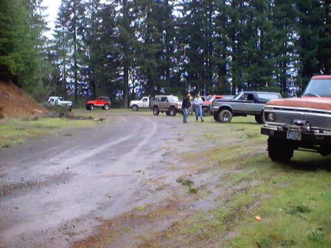 1976 Ford F-100 - A gathering of off-road vehicles on a wet, unpaved road surrounded by trees.