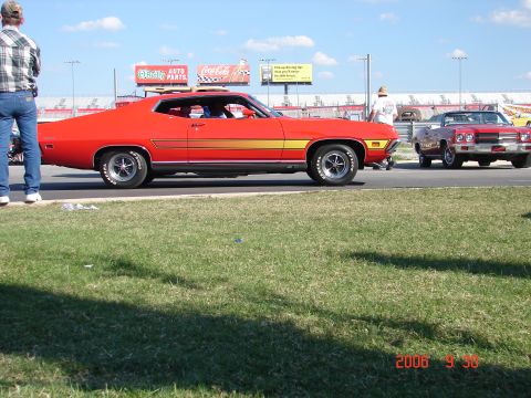1987 Ford Bronco - A classic red muscle car with a yellow stripe is parked at a racetrack, with another car visible in the background.