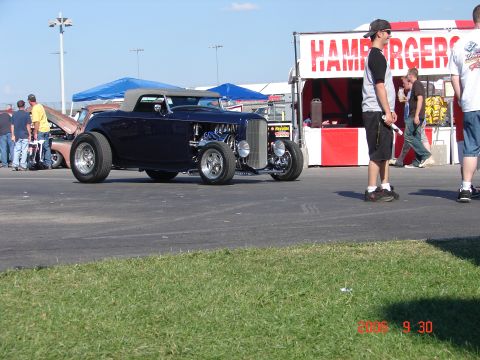 1987 Ford Bronco - A dark blue hot rod roadster is parked at an outdoor event, with people and a food stand in the background.