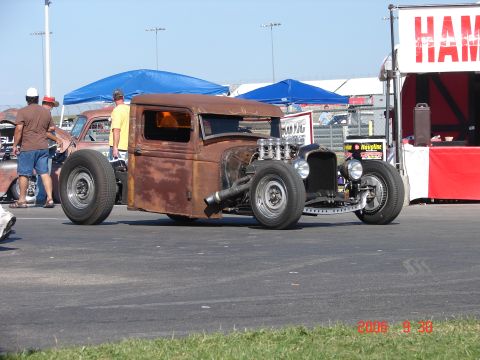 1987 Ford Bronco - A weathered hot rod pickup truck with a prominent V8 engine sits on pavement, with other vehicles and tents in the background.