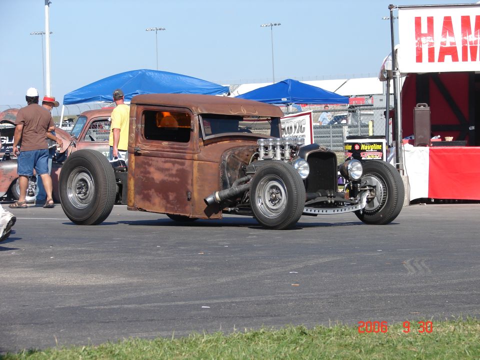 Rusty pickup truck with exposed engine and large rear tires. Another car is visible behind it