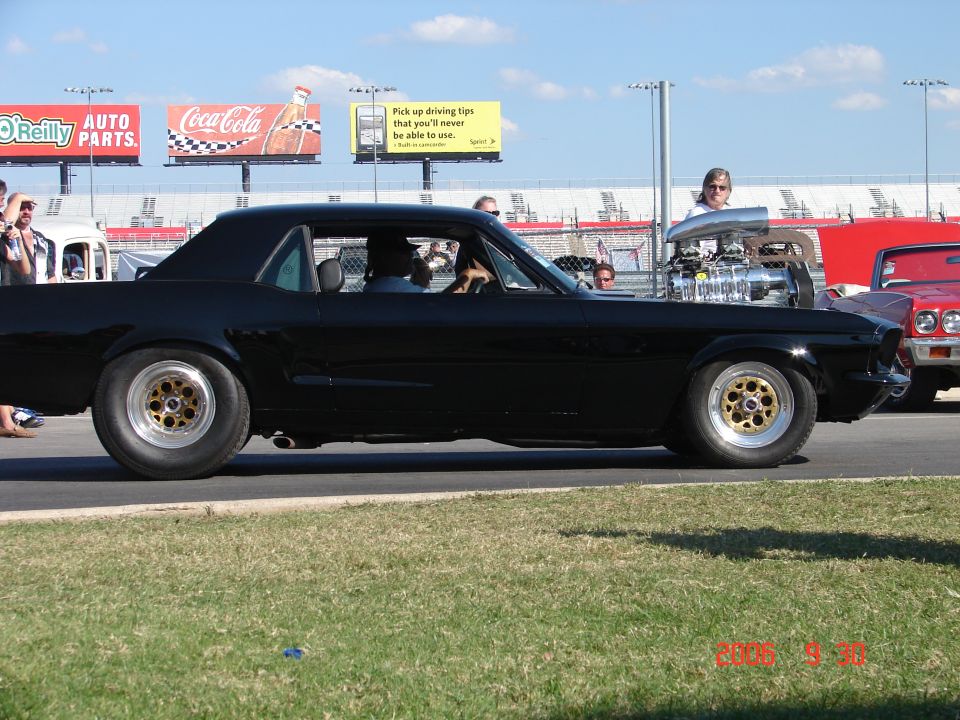 Black custom coupe with large supercharger protruding from hood, side view. Another car is visible to the right