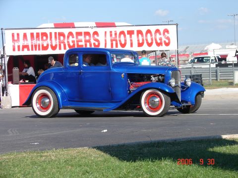 1987 Ford Bronco - A blue hot rod coupe with whitewall tires drives past a food stand with "HAMBURGERS | HOT DOGS" written on it.